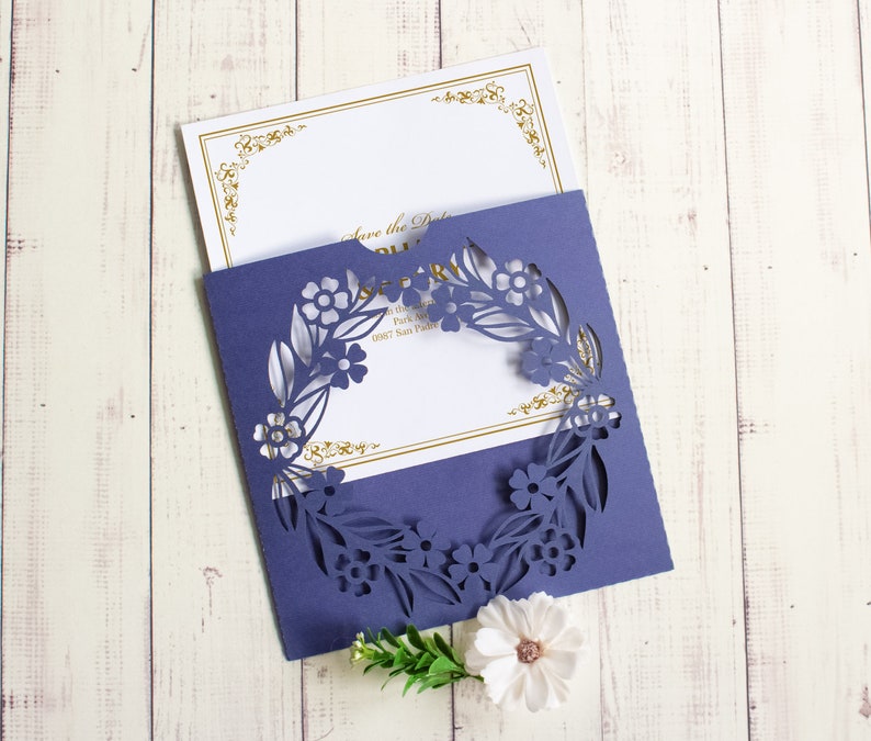 May include: A purple invitation card with a laser cut floral design. The card is folded in half and the front has a white insert with gold lettering that says "Save the Date".