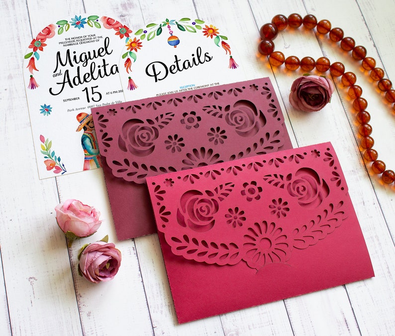 Puede incluir: Dos sobres de papel burdeos con intrincados dise&ntilde;os recortados que representan flores y hojas. Los sobres est&aacute;n sobre una superficie de madera blanca. El texto en el sobre dice "Detalles".