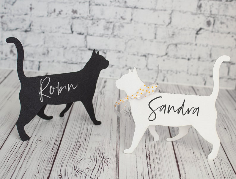 K&ouml;nnte beinhalten: Zwei schwarze und wei&szlig;e katzenf&ouml;rmige Platzkartehalter mit den Namen Robin und Sandra darauf geschrieben. Die Katze links ist schwarz und die Katze rechts ist wei&szlig;.