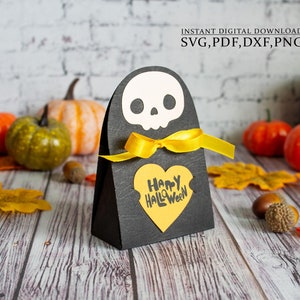 Könnte beinhalten: Eine schwarze Geschenkschachtel in Form eines Gespenstes mit einem weißen Schädel und einem gelben Band. Die Schachtel hat ein gelbes herzförmiges Etikett mit dem Text "Happy Halloween" darauf.