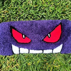 May include: A rectangular purple rug featuring a cartoon face with red eyes, white teeth, and a black outline. The rug is placed on a bed of green grass.