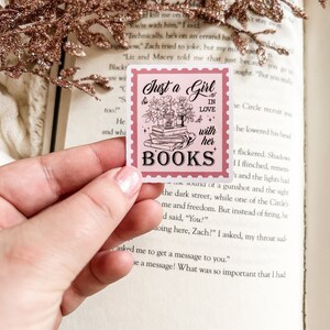 May include: A pink square sticker with a postage stamp design. The sticker reads "Just a Girl in Love with her Books" with floral and book illustrations. The sticker is held in a hand, with a book and decorative sprigs in the background.