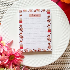 Peut inclure: Un bloc-notes blanc avec une bordure sur le thème de l'automne représentant des citrouilles, des tasses de café et des feuilles. Le bloc-notes a du papier ligné et le mot "Notes" imprimé en haut.
