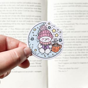 May include: A sticker featuring a cartoon snowman wearing a pink hat and scarf, holding a cup of hot chocolate and a book, sitting on a crescent moon with stars and snowflakes around it.