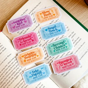 May include: A collection of colorful ticket-shaped stickers with various phrases like "Forbidden Love Book Club" and "Friends to Lovers Book Club." Each sticker has a unique color scheme and small illustrations.