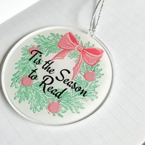 May include: White round ornament with a green wreath and a pink bow. The wreath has red ornaments and the text "Tis the Season to Read" in black.