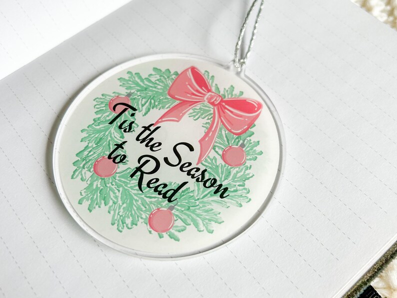 May include: White round ornament with a green wreath and a pink bow. The wreath has red ornaments and the text "Tis the Season to Read" in black.