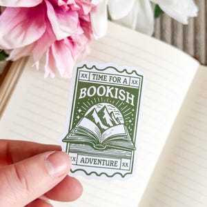 Peut inclure: Un autocollant vert et blanc avec le texte "Time for a Bookish Adventure" et une image stylisée d'un livre avec des montagnes en arrière-plan.
