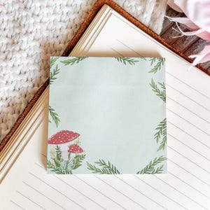 May include: A light blue sticky note with a green leafy border and a red and white mushroom illustration. The sticky note is on a lined notebook.