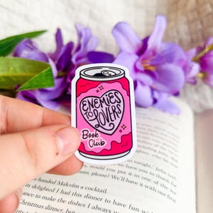 May include: A pink sticker in the shape of a soda can with the words "Enemies to Lovers Book Club" in a heart-shaped design. The sticker is held in front of a book and purple flowers.