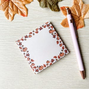 Könnte beinhalten: Ein quadratischer Notizblock mit einem Rand aus Kürbissen, Blättern und Eicheln in Orange und Braun. Ein hellrosa Stift mit goldener Spitze liegt auf einer hellen Holzoberfläche in der Nähe von Herbstblättern.