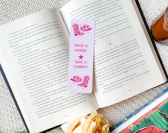 May include: A light purple bookmark with the text "SAVE A HORSE RIDE A COWBOY" and illustrations of cowboy boots and a hat. The bookmark is on top of an open book.