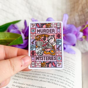 Könnte beinhalten: Ein rechteckiger Aufkleber mit den Worten "MURDER" und "MYSTERIES" in einem farbenfrohen, illustrierten Design. Das Design umfasst einen Schädel, ein Messer, Handschellen und andere kriminelle Symbole. Der Aufkleber wird vor einem Buch und lila Blumen gehalten.