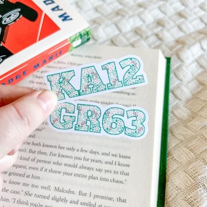 May include: Two stickers with the text "KA12" and "GR63" in a green font with a floral pattern. The stickers are on a book with a green spine. A book with a red cover is in the background.