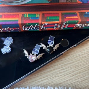 May include: Two heart-shaped earrings with bat wings, one pink and one black, with clear earring backs. The earrings are silver-toned and are on a reflective black surface. The book in the background reads "Eve's a Witch".