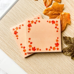 May include: Two square, peach-coloured notepads with a border of red and orange autumn leaves. The notepads are on a light wood-grain surface, with several decorative fall leaves scattered around.