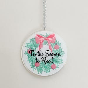 May include: A round, white Christmas ornament with a green and pink wreath and the text "Tis the Season to Read".