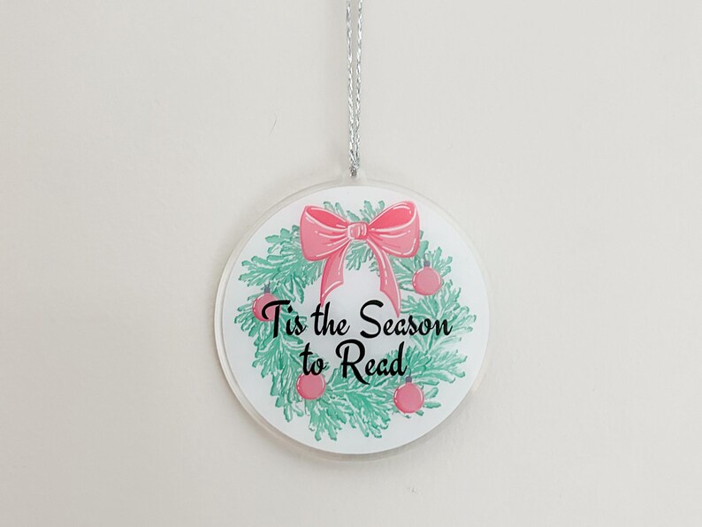 May include: A round, white Christmas ornament with a green and pink wreath and the text "Tis the Season to Read".