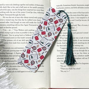 May include: A white and red bookmark with a Christmas theme. The bookmark features illustrations of books, candy canes, gingerbread cookies, and Christmas trees. The bookmark has a green tassel.