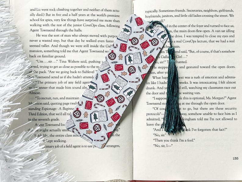 May include: A white and red bookmark with a Christmas theme. The bookmark features illustrations of books, candy canes, gingerbread cookies, and Christmas trees. The bookmark has a green tassel.