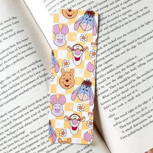 May include: A yellow and white checkered bookmark with cartoon characters from Disney's Winnie the Pooh and Disney's Winnie the Pooh. The bookmark features Winnie the Pooh, Tigger, Piglet, Eeyore, and a checkered background.