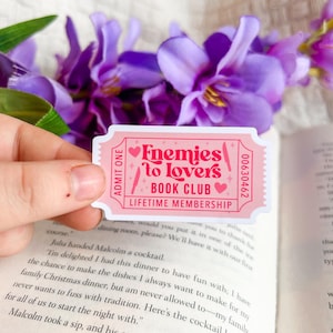 May include: A pink ticket-shaped sticker with the text "Enemies to Lovers Book Club Lifetime Membership" in a playful font. The sticker has a heart and knife design. The background includes purple flowers and an open book.