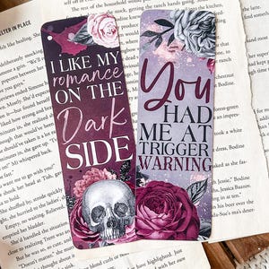 May include: Two purple and black bookmarks with floral designs. The first bookmark has a skull and the text "I like my romance on the dark side". The second bookmark has a rose and the text "You had me at trigger warning".
