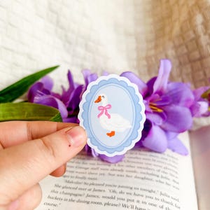 May include: A sticker of a white goose with orange feet and beak, wearing a pink bow, within a light blue oval frame with a scalloped edge. The sticker is held in front of a book and purple flowers.