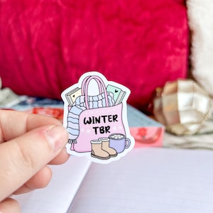 May include: A pink sticker featuring a tote bag filled with books, a scarf, and a sweater. The bag has the text "WINTER TBR" on it. Also included are a pair of boots and a mug of hot chocolate.