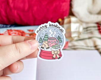 Może przedstawiać: Naklejka z motywem kuli śnieżnej z napisem "merry bookmas". Wewnątrz kuli znajdują się drzewa, kubek gorącej czekolady i szalik. Podstawa kuli jest różowa, a naklejka trzymana jest w dłoni.