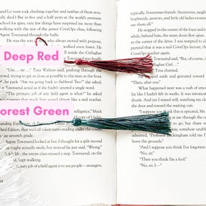 May include: A close-up of two book pages with a deep red tassel bookmark and a forest green tassel bookmark. The text on the pages is about a spy school and the students' experiences.