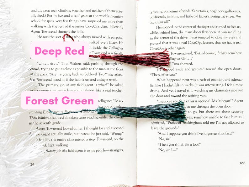 May include: A close-up of two book pages with a deep red tassel bookmark and a forest green tassel bookmark. The text on the pages is about a spy school and the students' experiences.