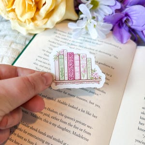 Peut inclure: Un autocollant représentant une étagère avec une collection de livres roses et verts, ornés de motifs floraux et décoratifs. L'autocollant est tenu au-dessus d'un livre ouvert, avec des fleurs en arrière-plan.