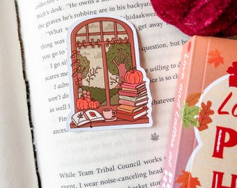 May include: A sticker featuring a window scene with pumpkins, books, and a cup of coffee. The window overlooks a tree and fall foliage. The sticker is on a book with the text "Love and Lattes at Pumphouse".