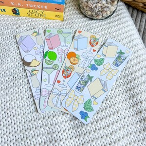 Könnte beinhalten: Vier dekorative Lesezeichen mit farbenfrohen Illustrationen von Cocktails, Büchern und Schleifen. Jedes Lesezeichen hat ein anderes Design, darunter eine Margarita, ein Orangencocktail und ein Getränk mit Blaubeeren und Minze. Die Lesezeichen liegen auf einer weißen Strickoberfläche.