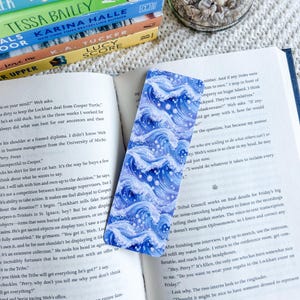 May include: A blue and white wave-patterned bookmark lies beside an open book. The bookmark has a repeating design of stylized ocean waves in shades of blue and white, creating a dynamic pattern. The bookmark is rectangular and appears to be made of paper.