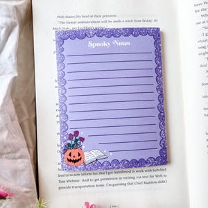 May include: A lilac notepad with the text "Spooky Notes" at the top. The notepad has a scalloped, glitter-edged border. A jack-o'-lantern, flowers, a spiderweb, and a book are in the bottom left corner.