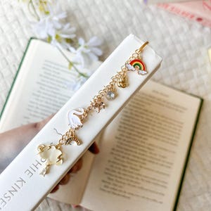 Könnte beinhalten: Ein goldfarbener Lesezeichen mit einer Kette aus Anhängern, darunter ein Regenbogen, ein Schwan und eine Wolke. Das Lesezeichen ist an einem Buch mit weißen Seiten befestigt. Die Anhänger sind in verschiedenen Farben gehalten, darunter Weiß, Gold und Regenbogenfarben.