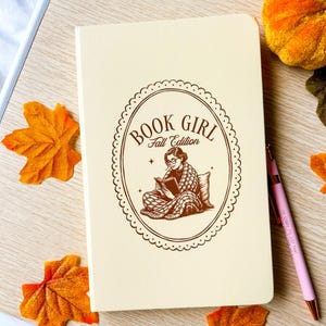 May include: Cream-coloured notebook with the text "BOOK GIRL Fall Edition" in a brown oval design. The design features a woman reading a book. A pink pen and autumn leaves are also visible.