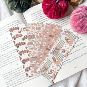 May include: A selection of decorative bookmarks with autumnal designs. The bookmarks display illustrations of trucks, books, coffee cups, and leaves in warm tones, ideal for readers.