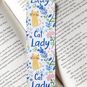 Peut inclure: Un marque-page blanc avec un motif floral et deux chats orange. Le marque-page porte le texte "Cat Lady" en lettres bleues répété trois fois.