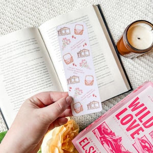 May include: A white bookmark featuring illustrations of a house, picnic basket, flowers, and strawberries. The text "IN MY SMALL TOWN ROMANCE ERA" is printed on the bookmark. The bookmark is positioned over an open book and a book titled "WILD LOVE".