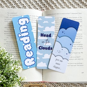 May include: Three bookmarks with cloud motifs. One features the word "Reading" in a bubble font, another says "Head in the Clouds," and the third says "just one more chapter..." in a blue gradient.