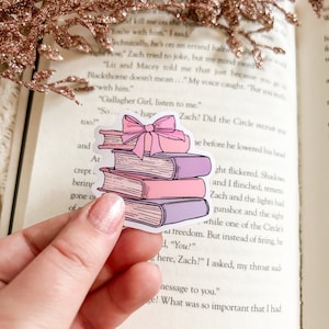 May include: A sticker of a stack of books with a pink bow on top. The books are pink, purple, and white with a light pink background.