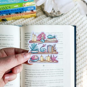 May include: A colourful sticker featuring a mermaid-themed bookshelf design. The sticker includes a mermaid, fish, books, candles, and starfish. The sticker is held in front of an open book.