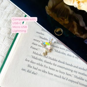 May include: A gold bookmark featuring a rainbow design and a dangling charm. The bookmark is placed on a book with text and a tablet. The image text reads "Compatible with: USB-C, Micro USB, Lightning."