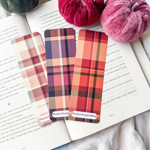 K&ouml;nnte beinhalten: Drei Lesezeichen mit Karomustern in Rot-, Rosa- und Lilat&ouml;nen mit abgerundeten Ecken werden auf einem offenen Buch pr&auml;sentiert. Die Lesezeichen tragen unten den Text "The Just Inspired Shop". Zwei Samtk&uuml;rbisse befinden sich im Hintergrund.