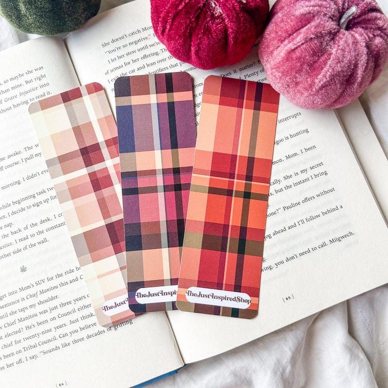K&ouml;nnte beinhalten: Drei Lesezeichen mit Karomustern in Rot-, Rosa- und Lilat&ouml;nen mit abgerundeten Ecken werden auf einem offenen Buch pr&auml;sentiert. Die Lesezeichen tragen unten den Text "The Just Inspired Shop". Zwei Samtk&uuml;rbisse befinden sich im Hintergrund.