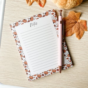 May include: A lined notepad with a pumpkin and leaf border, the word "Notes" in script, and a pink pen. The notepad and pen rest on a light wood surface, with fall leaves in the background. The notepad is approximately 15 cm long.