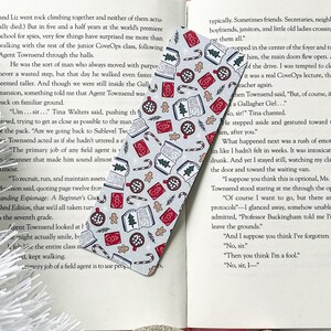 May include: A white and gray bookmark with a festive Christmas pattern. The bookmark features illustrations of gingerbread cookies, candy canes, mugs of hot chocolate, and books. The pattern is repeated throughout the bookmark.
